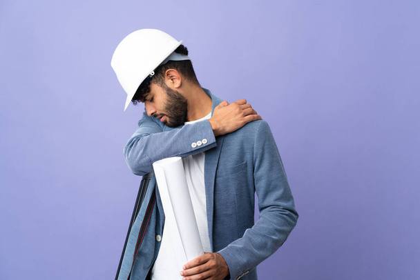 Young architect Moroccan man with helmet and holding blueprints over isolated background suffering from pain in shoulder for having made an effort - Foto, Imagem