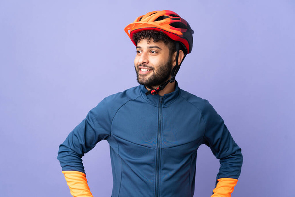 Νεαρός Μαροκινός ποδηλάτης απομονωμένος σε μωβ φόντο ποζάροντας με τα χέρια στο ισχίο και χαμογελώντας - Φωτογραφία, εικόνα