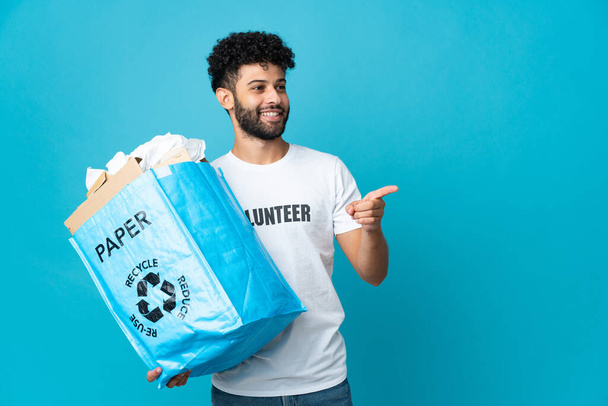 Young Moroccan man holding a recycling bag full of paper to recycle over isolated background pointing finger to the side and presenting a product - Photo, Image