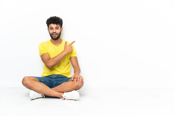 Young Moroccan handsome man sitting on the floor over isolated background surprised and pointing side - Photo, Image