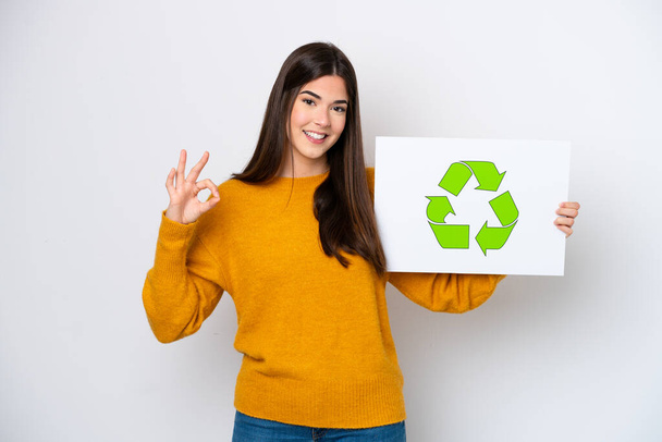 Young Brazilian woman isolated on white background holding a placard with recycle icon and celebrating a victory - Fotó, kép