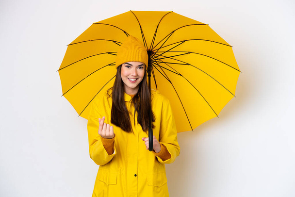Giovane donna brasiliana con impermeabile e ombrello isolato su sfondo bianco facendo gesto di soldi - Foto, immagini