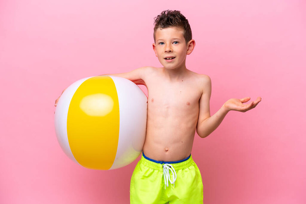 Pequeño chico caucásico sosteniendo una pelota de playa aislada sobre fondo rosa con expresión facial impactada - Foto, imagen