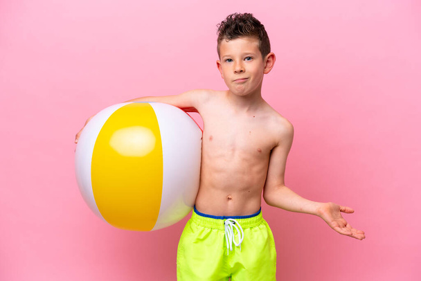 Маленький кавказский мальчик держит пляжный мяч, изолированный на розовом фоне, делая сомнительные жесты, поднимая плечи - Фото, изображение