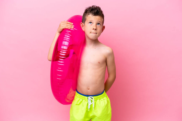 Piccolo ragazzo caucasico che tiene una ciambella gonfiabile isolata su sfondo rosa e alza lo sguardo - Foto, immagini
