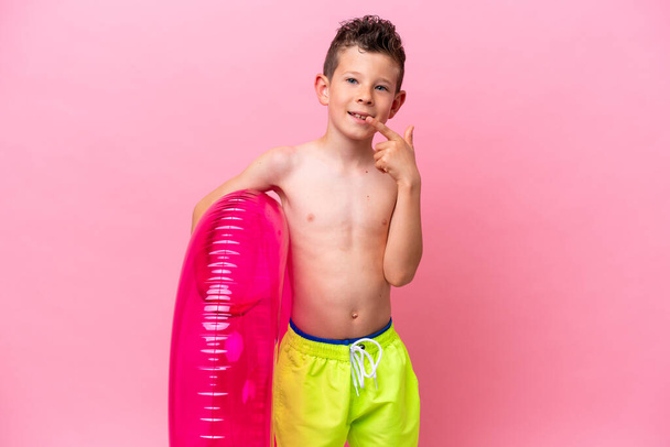 Маленький белый мальчик держит надувной пончик на розовом фоне, показывая большой палец вверх. - Фото, изображение