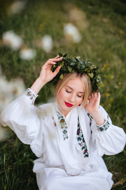 A Ukrainian woman in a white embroidered shirt and with a wreath on her head in a blooming garden in spring. Portrait of a Slavic girl. - Foto, Bild