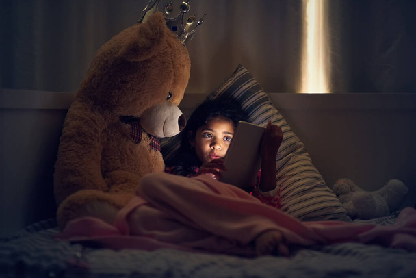 Kijken naar haar favoriete tekenfilms voor het slapen gaan. Shot van een klein meisje met behulp van een digitale tablet terwijl liggend in bed met haar teddy 's nachts. - Foto, afbeelding