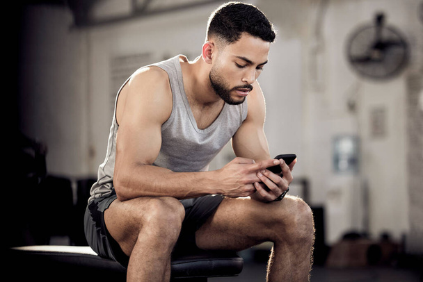 Перерыв означает обновление спортзала. Снимок мускулистого молодого человека, использующего мобильный телефон в спортзале. - Фото, изображение