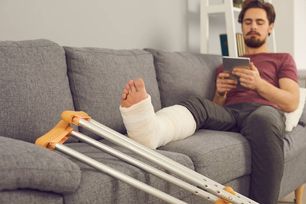 Young man sitting on sofa with broken leg in cast - Foto, Bild