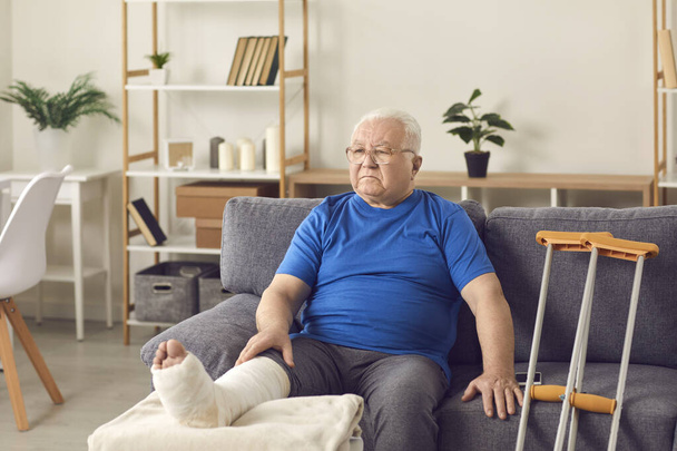 Disappointed senior mature man sitting on sofa with broken leg in cast - 写真・画像