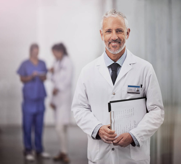 Vaše zdraví je to, pro co pracuji. Portrét dospělého mužského lékaře stojícího v nemocnici se svými kolegy v pozadí. - Fotografie, Obrázek