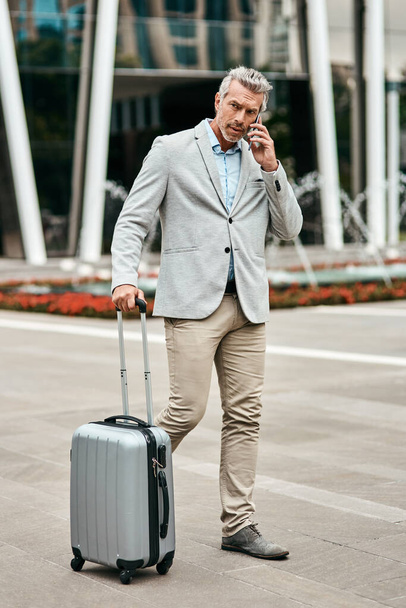 Şehri ele geçirmek için hazır. Yetişkin bir iş adamının şehirde bavulla yürürken cep telefonuyla konuşması.. - Fotoğraf, Görsel