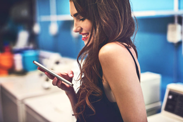 Планирую с друзьями. Фотография привлекательной молодой женщины, которая пишет смс на телефоне, пока стирает вещи в прачечной днем.. - Фото, изображение