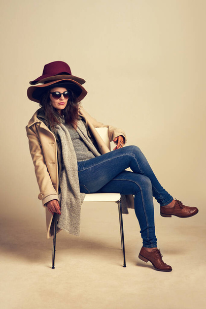 Complétez votre tenue avec le chapeau parfait. Plan studio d'une jeune femme assise sur une chaise et portant une pile de chapeaux sur un fond brun. - Photo, image