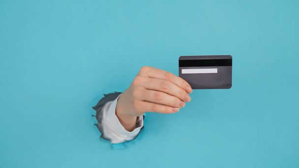 A womans hand sticking out of a hole in a paper blue background holds a credit card.  - Foto, Imagem