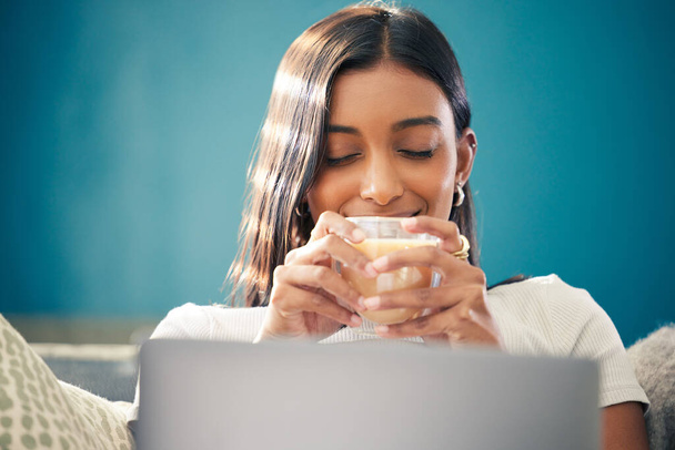 Hmm, nothing like a hot cup of tea. Shot of a young woman sitting in her living room and enjoying a cup of tea while using her laptop. - Foto, imagen