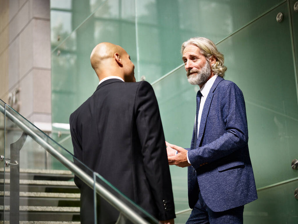 caucasian and latino corporate business men talking chatting having a conversation while walking on stairs in modern office building - 写真・画像