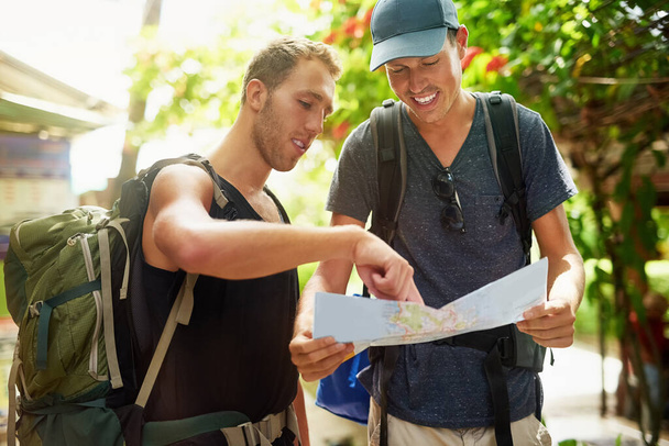 新しい地区への道を見つけること。タイにいる間に地図上で一緒に話をするバックパックを着ている2人の笑顔の若い友人のショット. - 写真・画像