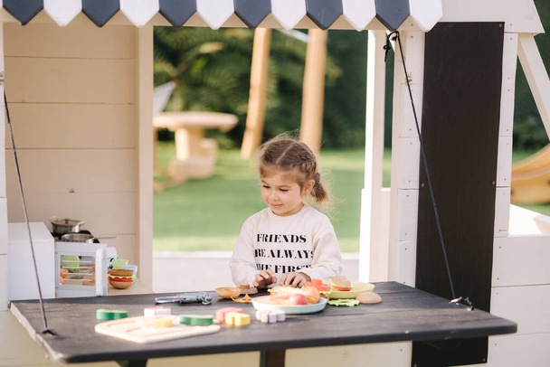 Happy little girl playing on toy kitchen on wheels. Cute girl make a burger on toy kitchen outdoors. Adorable kid play on fresh air - Foto, Imagen