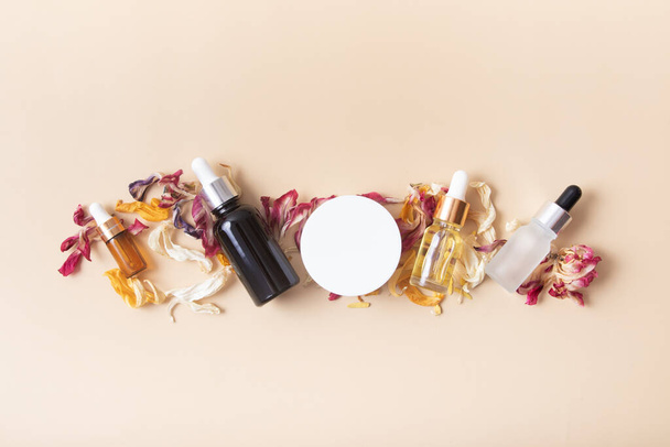 Botellas cosméticas y frasco con pétalos de flores de color seco en una vista superior de fondo beige. - Foto, imagen
