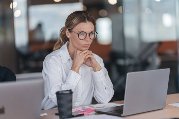 Mujer de negocios seria con gafas que trabajan en el ordenador portátil en el lugar de trabajo en la oficina moderna - Foto, Imagen