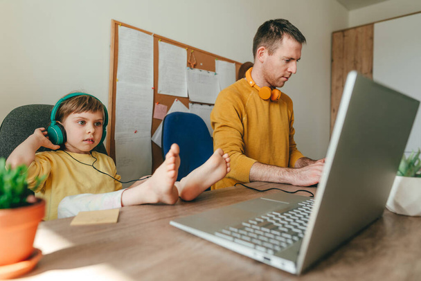 Egy 5 éves kislány az apjával, aki laptopot használ az asztalnál ülve. A család technológiát használ otthon. Szülőnek lenni a kovid-19-es zárlat alatt. Apa és a lánya otthon dolgoznak. Szelektív fókusz. - Fotó, kép