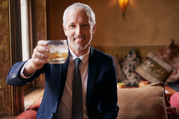 Raise a glass with me.... Portrait of a well-dressed mature man making a toast with a glass of whiskey in a bar after work. - Zdjęcie, obraz