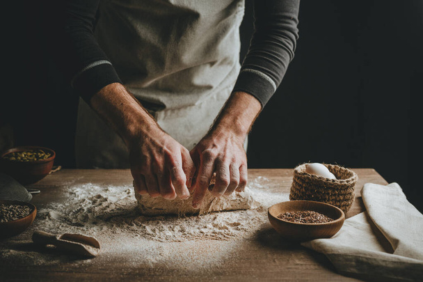 Unrecognizable young man kneading dough on wooden table. Males hands making bread on dark background. Selective focus. - Fotoğraf, Görsel