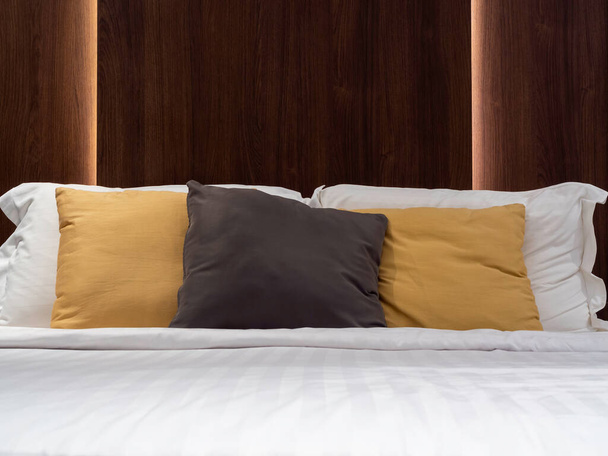 Set di cuscini puliti, marrone, giallo e bianco, coperta e lenzuolo nella camera d'albergo con testiera in legno scuro e luce dietro di essa. Lenzuola in camera da letto, vista frontale. - Foto, immagini