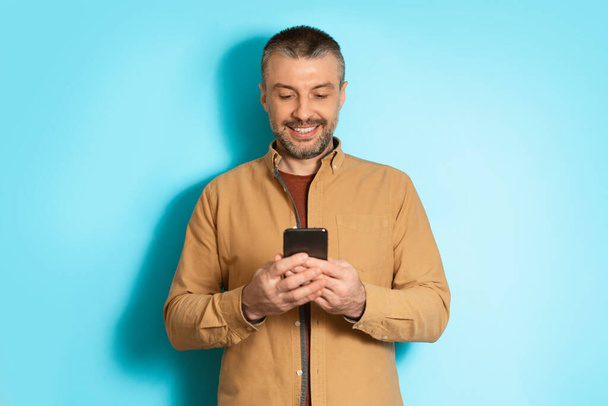 Χαρούμενος άνθρωπος που χρησιμοποιεί το κινητό εφαρμογή στο κινητό τηλέφωνο πάνω από το μπλε φόντο - Φωτογραφία, εικόνα
