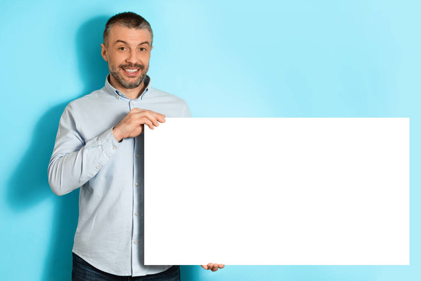 чоловік середнього віку показує порожню дошку паперу на блакитному тлі
 - Фото, зображення