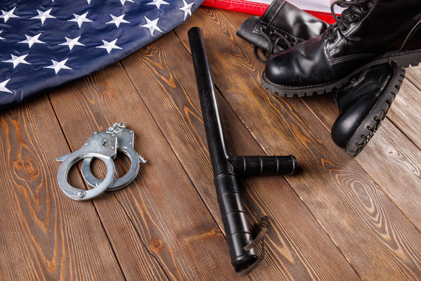 silver metal handcuffs, black ankle boots and police nightstick near US flag on wooden surface - Foto, imagen