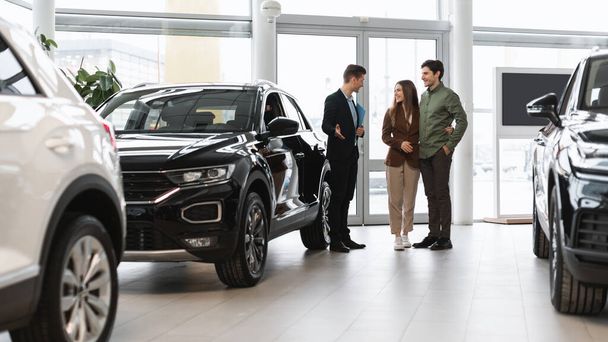 Car purchase or rental. Positive young spouses speaking to salesman about buying new auto at dealership, panorama - Fotografie, Obrázek