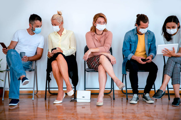 Pessoas com máscara facial sentadas em cadeiras seguidas. Grupo de pessoas multiétnicas em sala de espera - Foto, Imagem