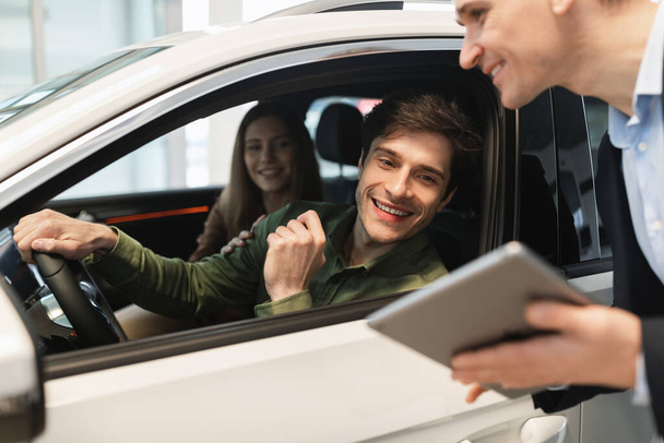 Young Caucasian couple clarifying car purchase details with salesman, looking at tablet screen in dealership center - Foto, afbeelding