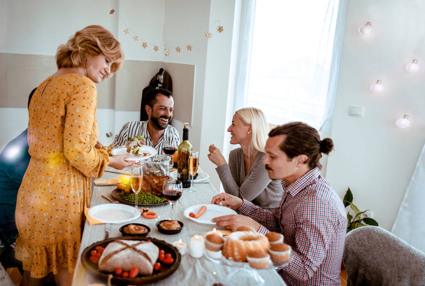 Weseli, wieloetniczni przyjaciele zjedzący świąteczną kolację, cieszący się świętowaniem świąt razem z jedzeniem i śmiechem. Rodzinne spotkanie przy stole kuchennym w Święto Dziękczynienia - Zdjęcie, obraz