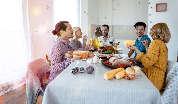 陽気な多民族の友人がお祝いの夕食を食べ、食べ物や笑いと一緒に休日を楽しんでいます。感謝祭の日にキッチンテーブルを囲んで家族の集まり - 写真・画像