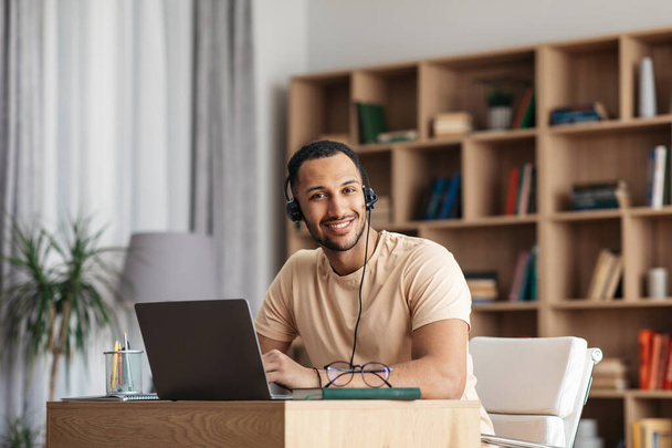 Portrait of excited arab man in headphones having online meeting on laptop and smiling at camera, working from home - Zdjęcie, obraz