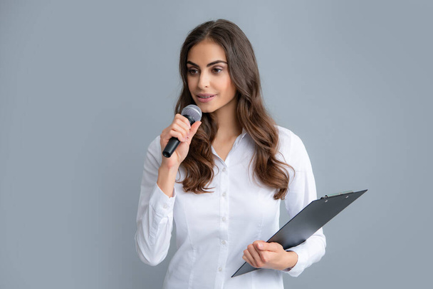 Портрет молодой улыбчивой деловой женщины, делающей презентацию с буфером обмена и микрофоном на изолированном сером фоне. - Фото, изображение