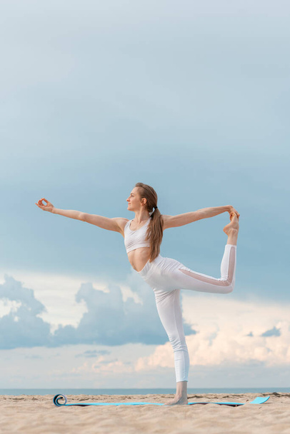 Young woman in white sportwear practicing yoga in the morning on seashore.Yoga at the beach Doing asana Natarajasana. - 写真・画像