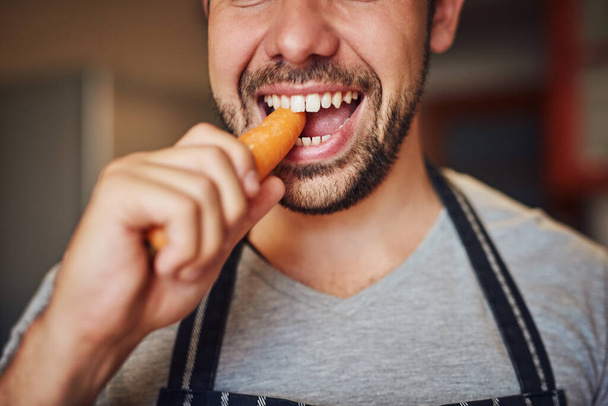 Hij heeft zin in caroteen. Gehakt van een onbekende jongeman die thuis een wortel eet in zijn keuken.. - Foto, afbeelding