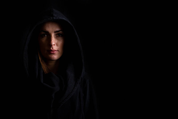 Sad young woman in a black hood on a dark black background. - Фото, изображение