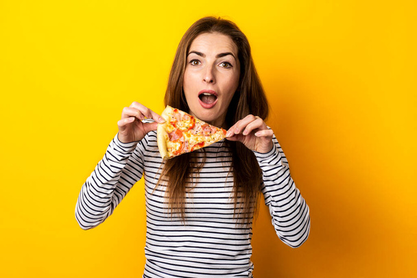 surprised shocked young woman looks holding a slice of hot fresh pizza on a yellow background. - Fotoğraf, Görsel