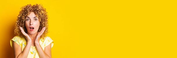 έκπληκτος σγουρά νεαρή γυναίκα κρατώντας παλάμες κοντά στο πρόσωπο σε κίτρινο φόντο. Μπάνερ. - Φωτογραφία, εικόνα