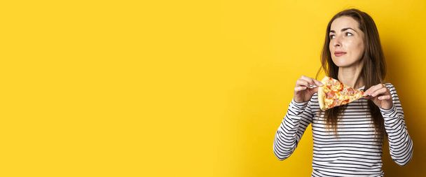 young woman looks to the side holding a slice of hot fresh pizza on a yellow background. Banner. - Fotoğraf, Görsel