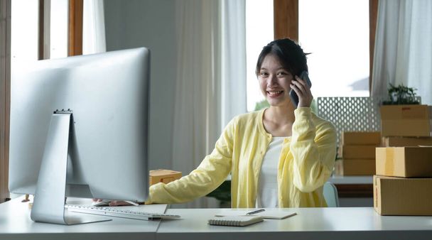 A portrait of Asian woman, e-commerce employee sitting in the office full of packages on the table using a laptop and smartphone, for SME business, e-commerce, technology and delivery business - Foto, Imagem