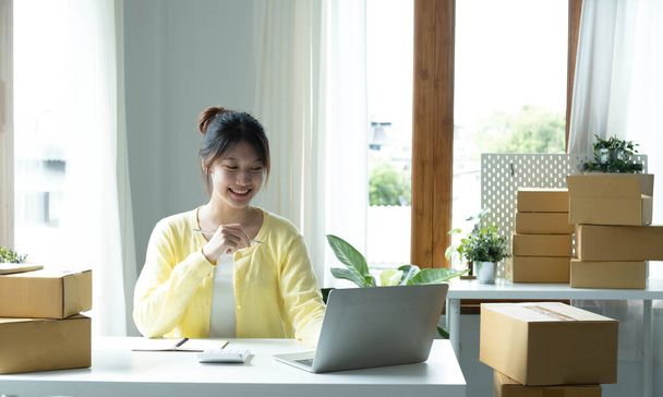 A portrait of Asian woman, e-commerce employee sitting in the office full of packages on the table using a laptop and calculator, for SME business, e-commerce, technology and delivery business - Foto, Imagem