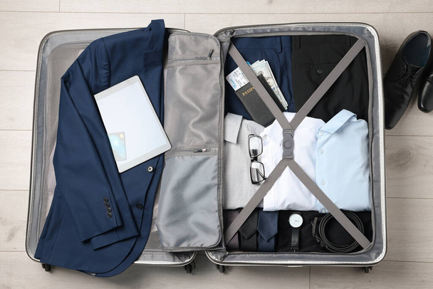 Folded clothes with accessories in open suitcase and shoes on wooden floor, flat lay. Business trip planning - Foto, Bild
