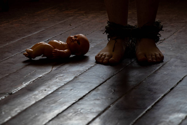 Mit einem Seil gefesselte Füße und eine Babypuppe auf einem Holzboden im Dunkeln, Menschen in Sklaverei, Frauenhandel - Foto, Bild